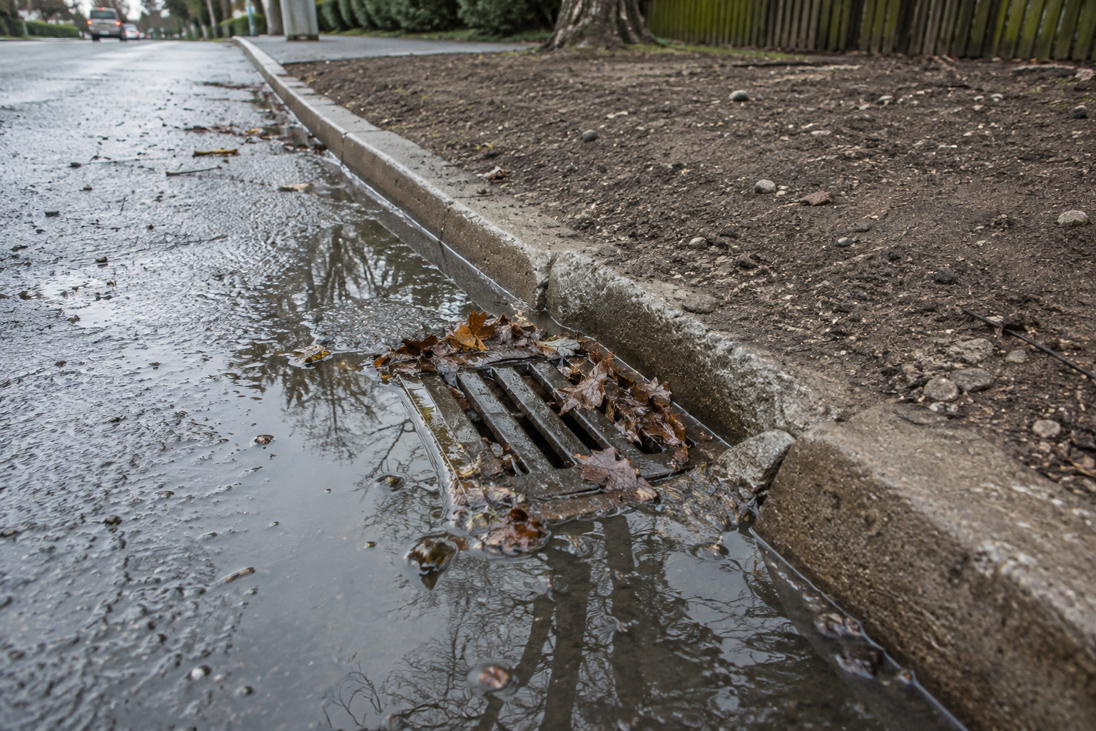 Surface water flooding beside a leaf-blocked roadside gully and dry soil verge.